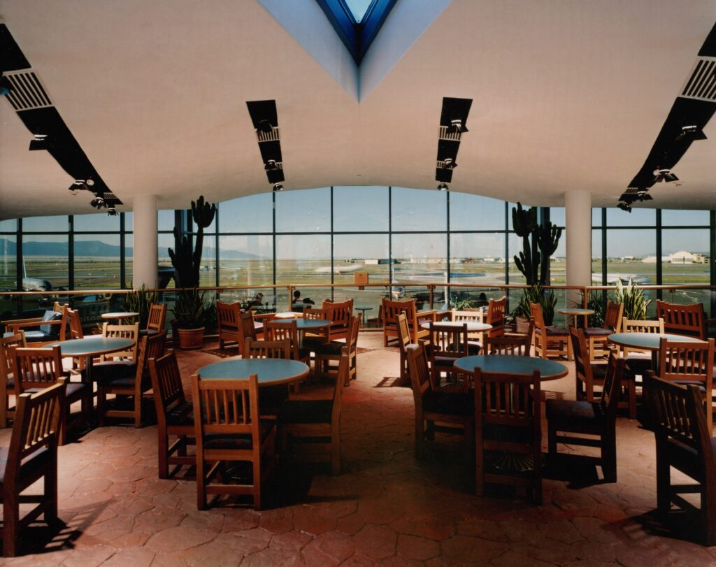 Bright, spacious airport lounge features wooden tables and chairs, with large windows overlooking the runway and desert landscape outside.