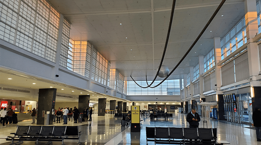 Spacious airport terminal interior with large windows and modern architecture. Passengers gather near check-in counters and seating areas.