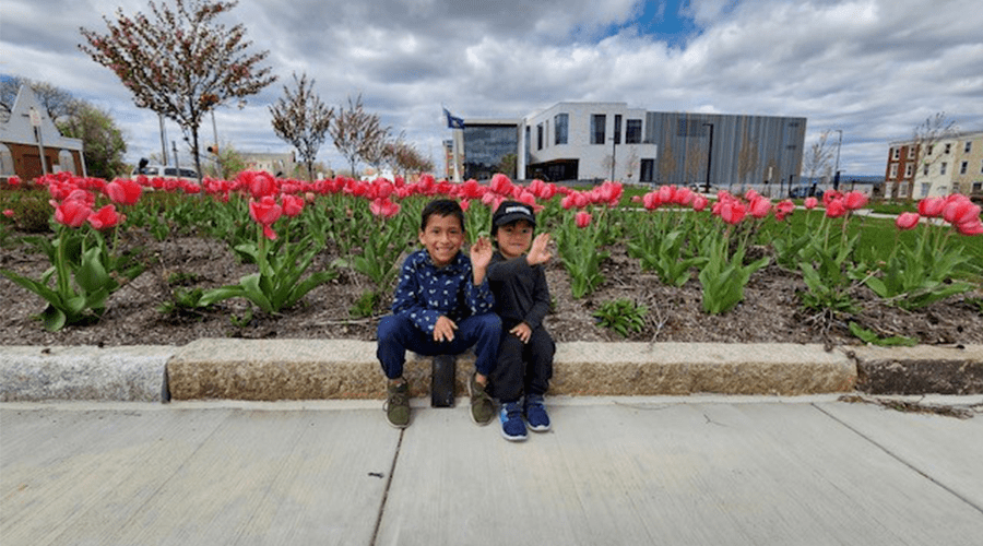 Two young boys sit on a stone edge, smiling and waving amidst vibrant pink tulips in a park, with a modern building in the background.