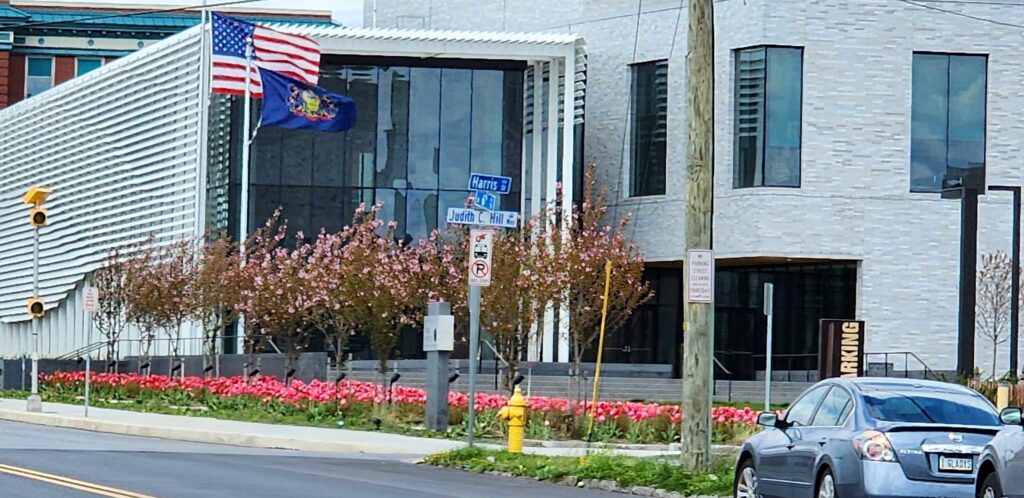 Modern building featuring American and Pennsylvania flags, surrounded by flowering plants. Street signs indicate Harris and Judith C. Hill streets.