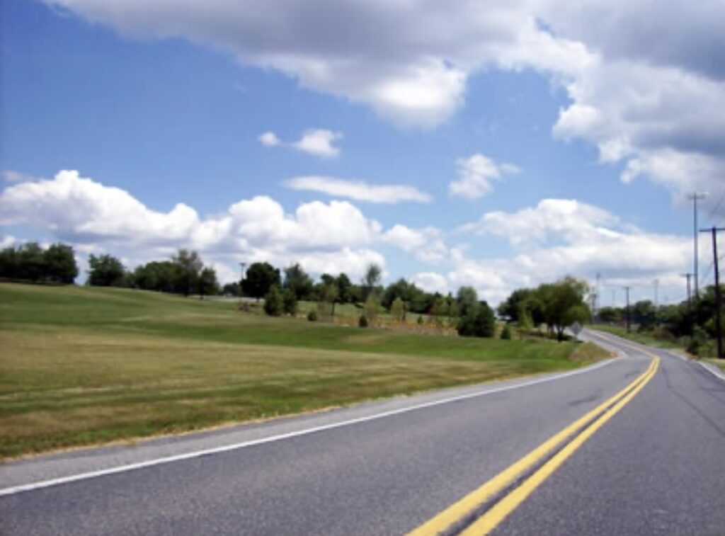 Curving rural road bordered by green fields and trees under a bright blue sky with clouds. Ideal for showcasing scenic driving routes.